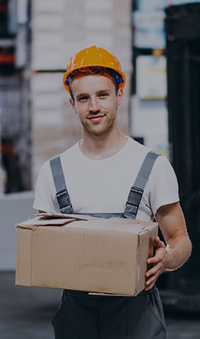 ceci est une photo d'un homme avec un casque de chantier, en t-shirt et salopette de travail, il tiens un carton